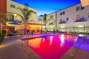 A swimming pool with a red-lit water feature, surrounded by lounge chairs and a bar area with string lights.
