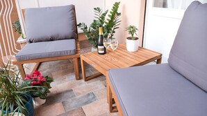 Un espace patio comprenant deux fauteuils gris avec coussins, une table en bois sur laquelle se trouvent une bouteille de champagne et deux verres, ainsi que diverses plantes en pot.