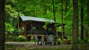 A cabin with a porch, two chairs, and a table in a forest. - Cozy Cabin Among the Trees - Rustic Charm, Modern Comforts,  26 Secluded Acres  (Stoystown)