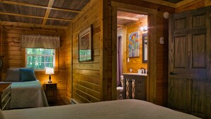 A wooden cabin bedroom with a bed, bedside table, lamp, and a view of the bathroom. - Cozy Cabin Among the Trees - Rustic Charm, Modern Comforts,  26 Secluded Acres  (Stoystown)