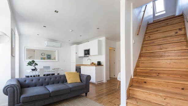 A modern living room with a grey sofa, a wooden staircase, and a kitchen in the background. - The Tin Artist Atelier 1 bedroom apartment (Lisboa)