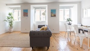 A spacious living area with a grey sofa, white chairs, a dining table, and a large window with a view of buildings. - The Tin Artist Atelier 1 bedroom apartment (Lisboa)