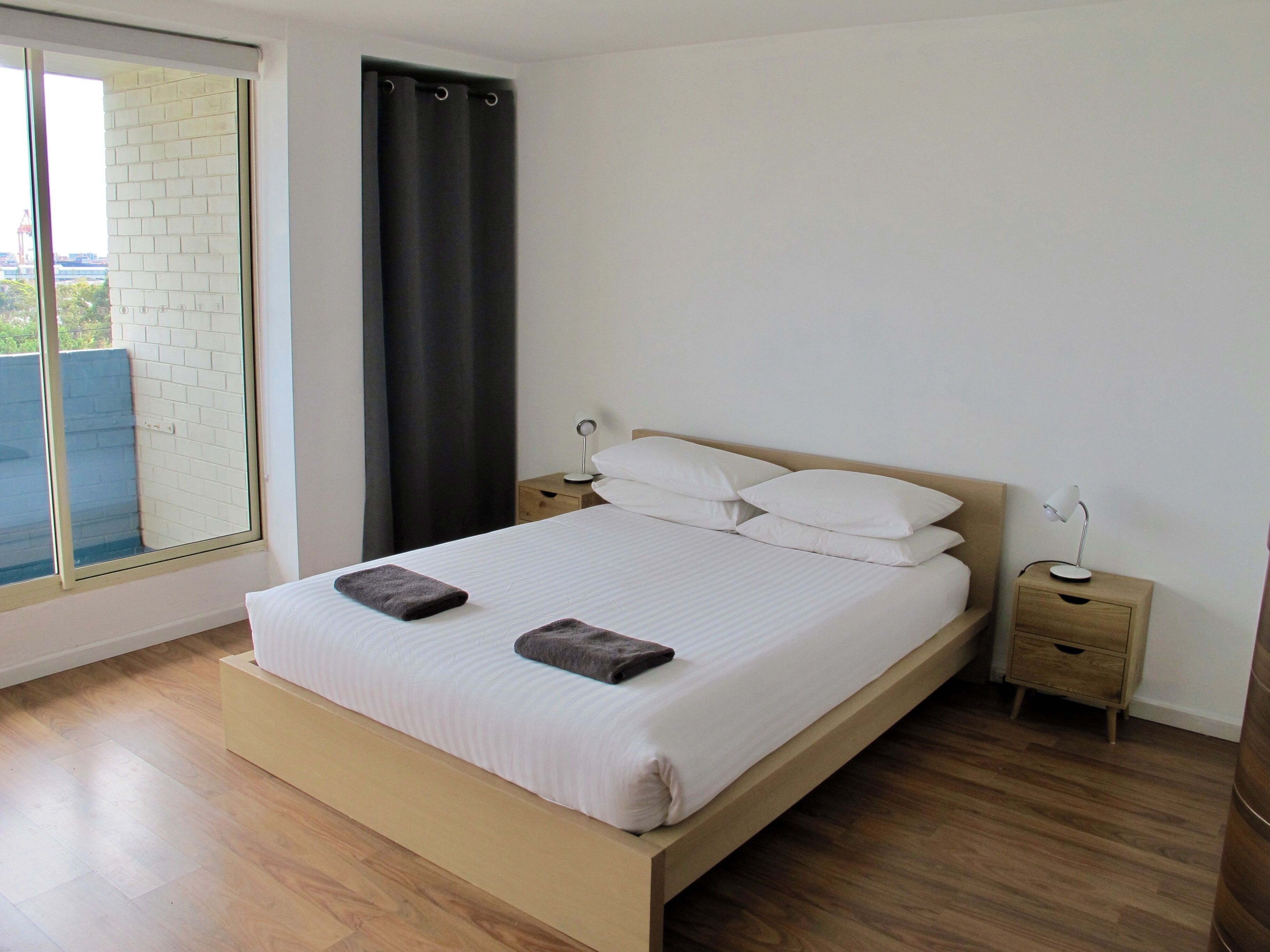 Une chambre à coucher avec un lit, deux tables de chevet, une fenêtre avec un rideau et une vue sur un mur de briques et de la verdure à l’extérieur.