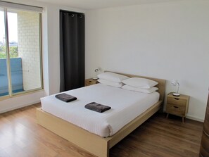A bedroom with a bed, two bedside tables, a window with a curtain, and a view of a brick wall and greenery outside.