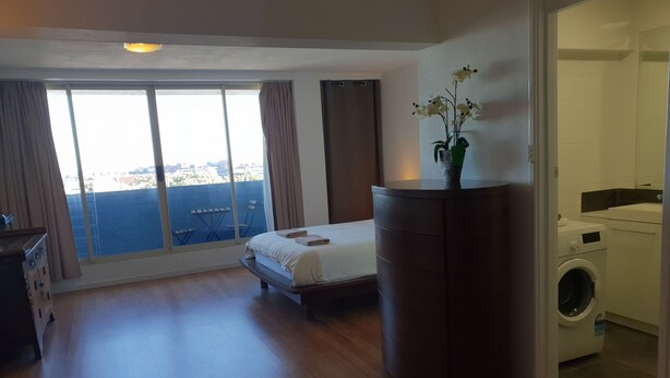 A hotel room with a bed, a small desk, a washing machine, and a view of the city through large windows. - Champagne views on a budget (Fremantle)