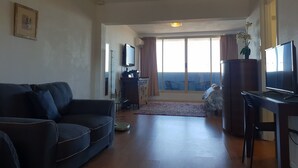 A living room with a sofa, a TV, a wooden cabinet, and a view of the outside through large windows. - Champagne views on a budget (Fremantle)