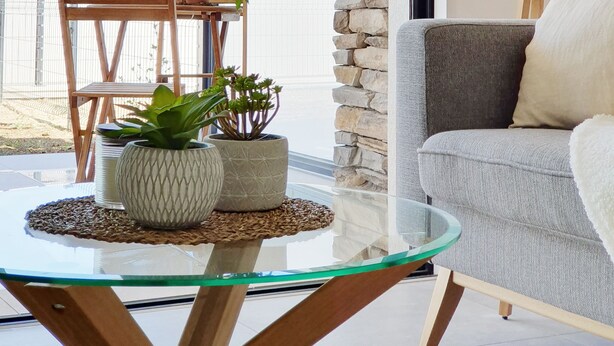 Una sala de estar moderna con una mesa de centro de vidrio, un sofá gris y una pared de acento de piedra. Hay una planta en maceta sobre la mesa y una silla de madera al fondo.