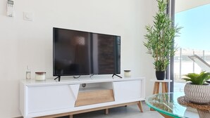 Una sala de estar moderna con un televisor de pantalla plana sobre un mueble blanco, una mesa de centro de vidrio y una planta en maceta.