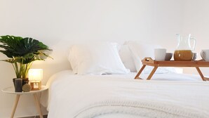 Una cama bien hecha con ropa blanca, una mesita de noche con lámpara, una planta en maceta y una bandeja con jarras y tazas.
