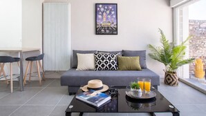 A modern living room with a grey sofa, a dining area with two chairs, a large window with a view of a brick wall, and a coffee table with a book and a tray of orange juice. - Modern apartment with 40m² terrace - Extremely practical (Arras)