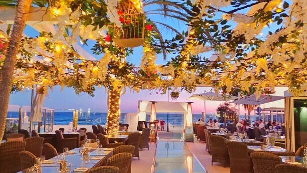 Un área de comedor al aire libre con mesas y sillas de mimbre, una piscina y vistas al mar.