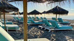 Una playa con filas de sillas reclinables turquesa y sombrillas de paja.