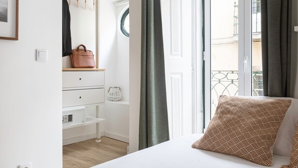 A bedroom with a bed, a dresser, a hanging coat rack, and a window with curtains. - The Iron Artist Studio Atelier (Lisboa)