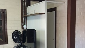 A room with a white desk, a black fan, and a television. There is a white brick wall and a wooden beam ceiling. - Casa do Sassá - Close to everything (Guarda do Embaú)