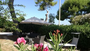 En udendørs terrasse med fletmøbler, en stor parasol og blomstrende planter.