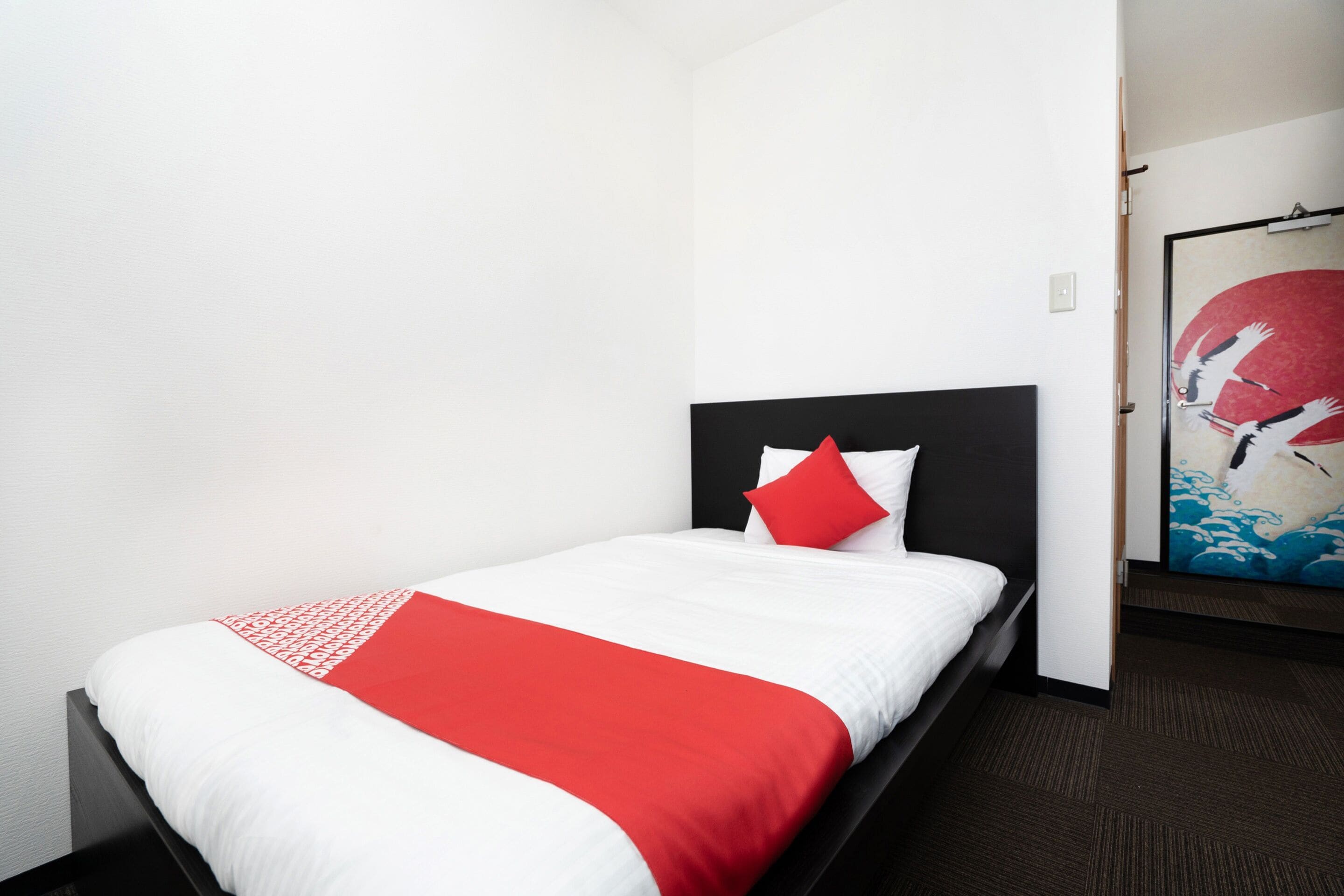 Quarto de hotel com uma cama de solteiro, edredom vermelho e branco e um quadro na parede.