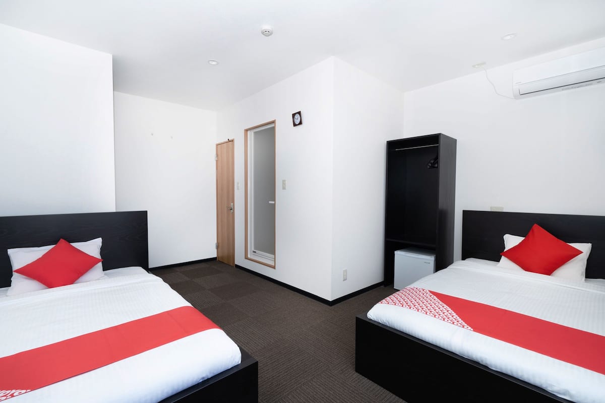 Habitación de hotel con dos camas, cada una con colcha roja y blanca, un armario y una puerta que da a otra habitación.