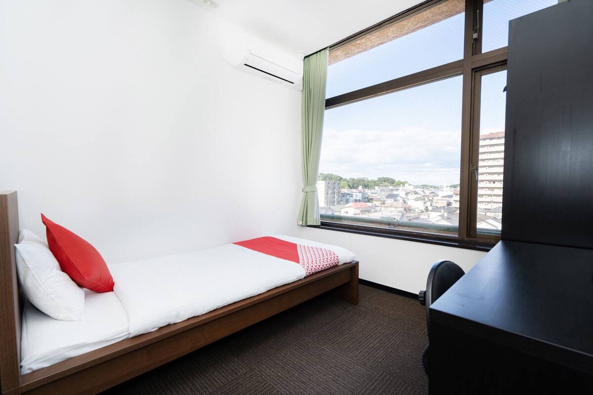 Una cama individual con ropa de cama roja y blanca, un escritorio y una ventana con vista a un paisaje urbano.