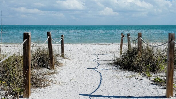 Un sentier de sable qui longe la plage et mène à l’océan, avec des poteaux en bois et des cordes qui délimitent l’entrée.