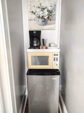 Cocina compacta con microondas y cafetera sobre una encimera, refrigerador debajo y una obra de arte floral enmarcada en la parte superior.