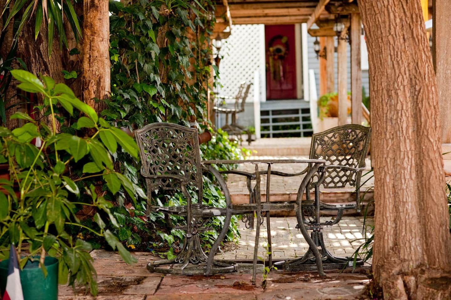 An outdoor seating area with wrought iron chairs and tables, surrounded by lush greenery and a wooden pergola.