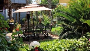 Eine Außenpatio-Terrasse mit Tisch und Stühlen unter einem Sonnensegel, umgeben von üppiger Bepflanzung und hohen Palmen.