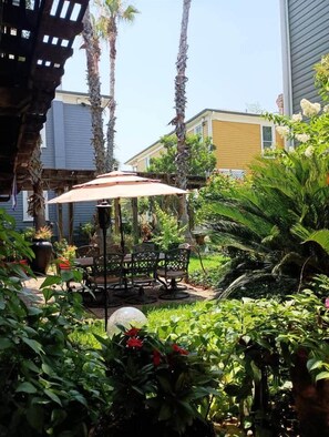An outdoor patio area with a table and chairs under a canopy, surrounded by lush greenery and tall palm trees.