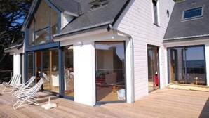 Una casa moderna con ventanas amplias, una terraza de madera y un portón de cristal que da al interior.