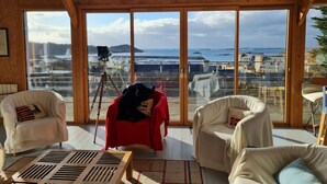 Amplia habitación con grandes ventanales que ofrecen vista al mar y a edificios. Hay dos sillas blancas, una mesa de madera y un trípode con una cámara.
