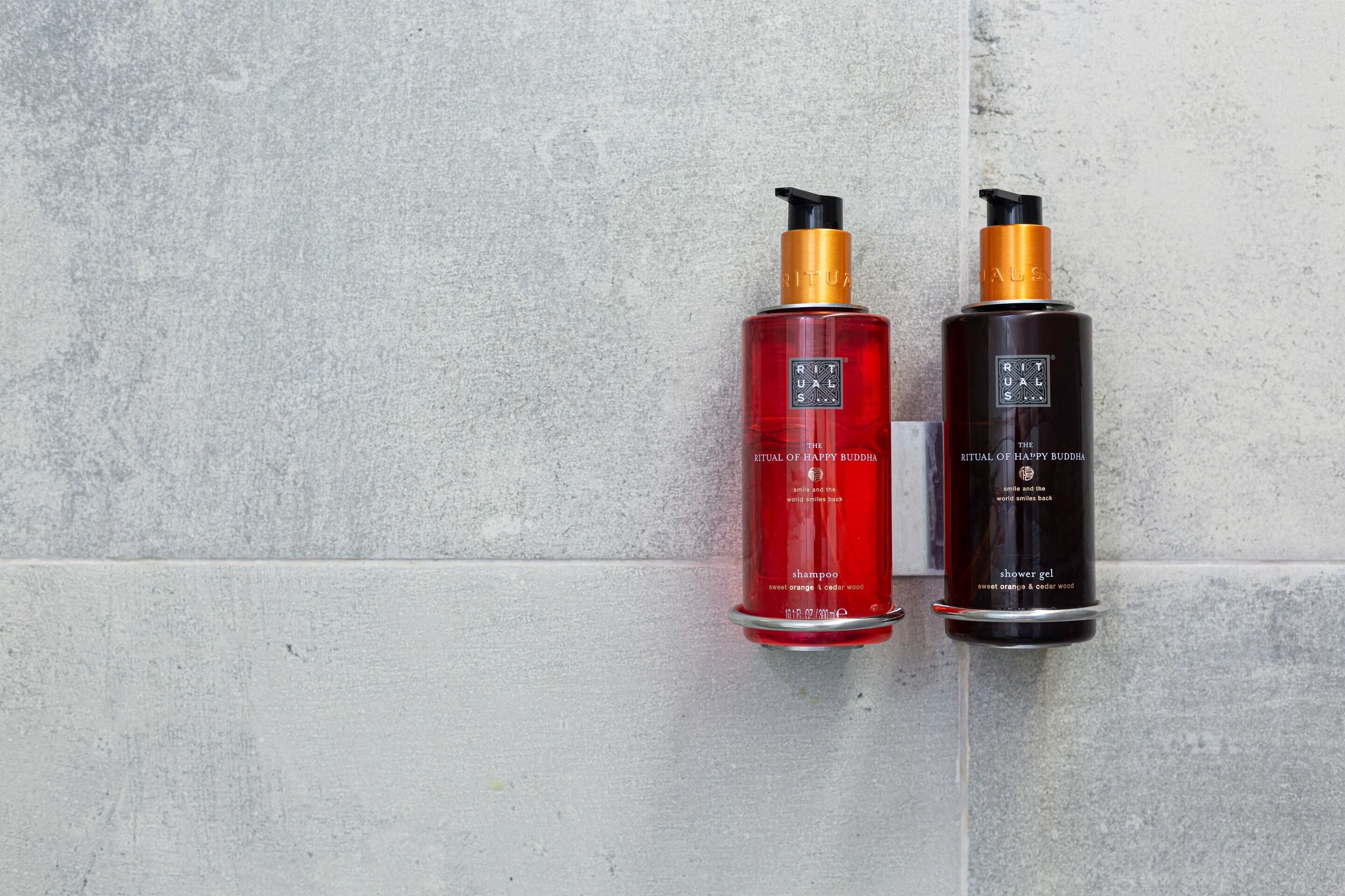 Deux bouteilles de shampoing et de gel douche de la gamme Ritual of Happy Buddha, posées devant un mur à texture.