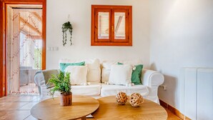 Un salón con un sofá blanco, dos mesas redondas de madera, una planta en maceta y una ventana con marcos de madera.