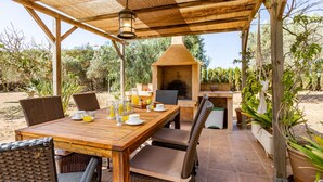 Un patio exterior con una mesa de madera dispuesta para el desayuno, sillas de mimbre y un horno de piedra.