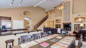 Amplia sala de estar con techo alto, una escalera central, una cocina con refrigerador de acero inoxidable y una zona de estar con una mesa grande y sillas.