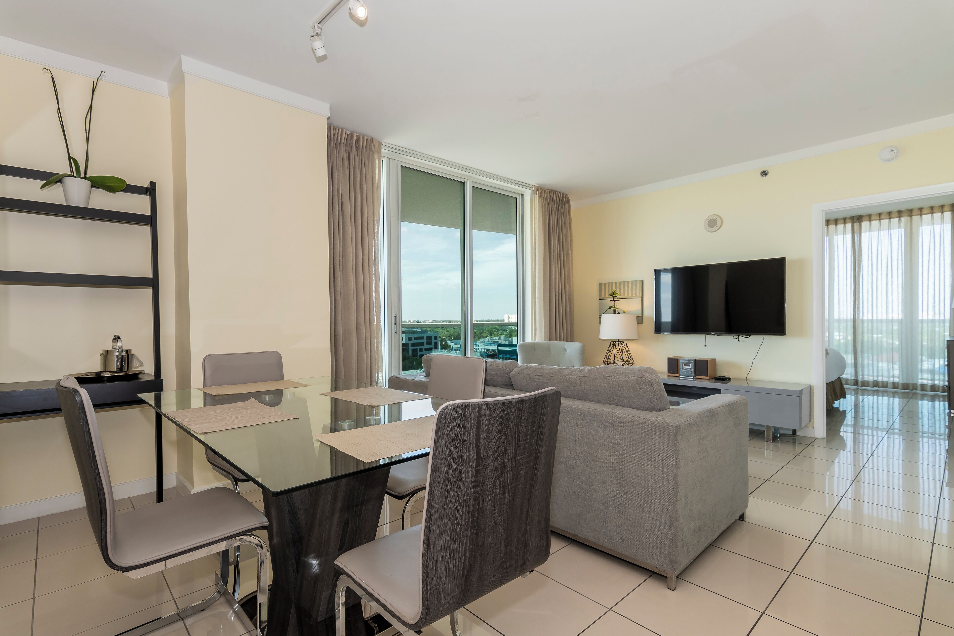 A modern living room with a glass dining table, grey sofas, and a flat-screen TV.