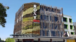Un edificio moderno con fachada de jardín vertical, un letrero llamativo que dice "ROXY PADUNGAN" y una entrada a nivel de calle con un anuncio de alquiler de coches.