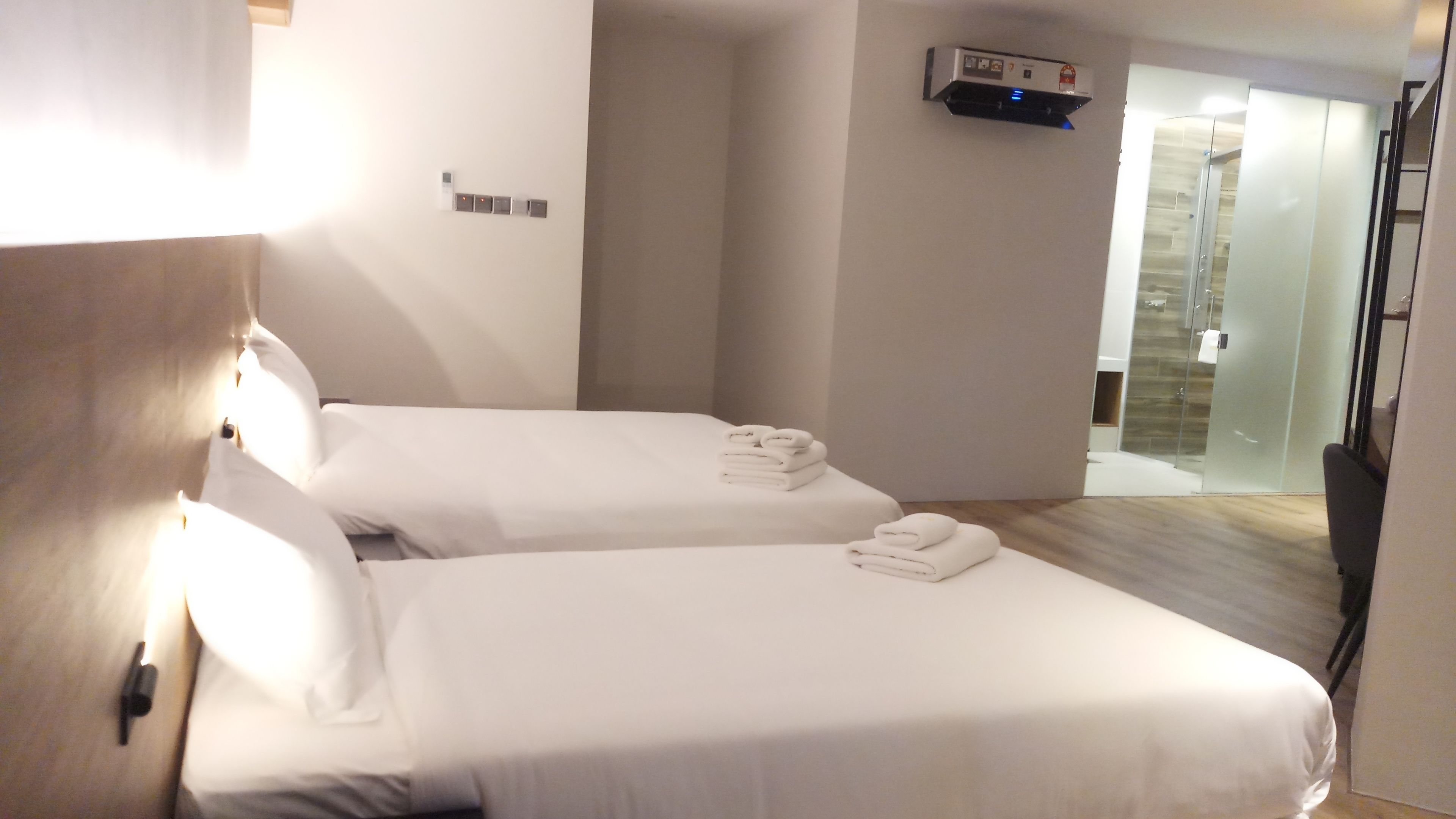 A hotel room with two single beds, a bathroom with a glass shower, and a mounted air conditioning unit.
