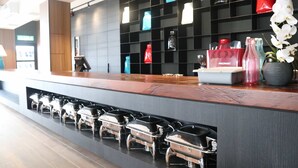 Una zona de recepción moderna de hotel con mostrador, máquina de café y varias estaciones tipo bufé.