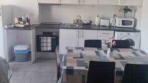 Una cucina compatta con mobili bianchi, un forno a microonde e un lavello. Si nota un tavolo con sedie e un cestino blu.