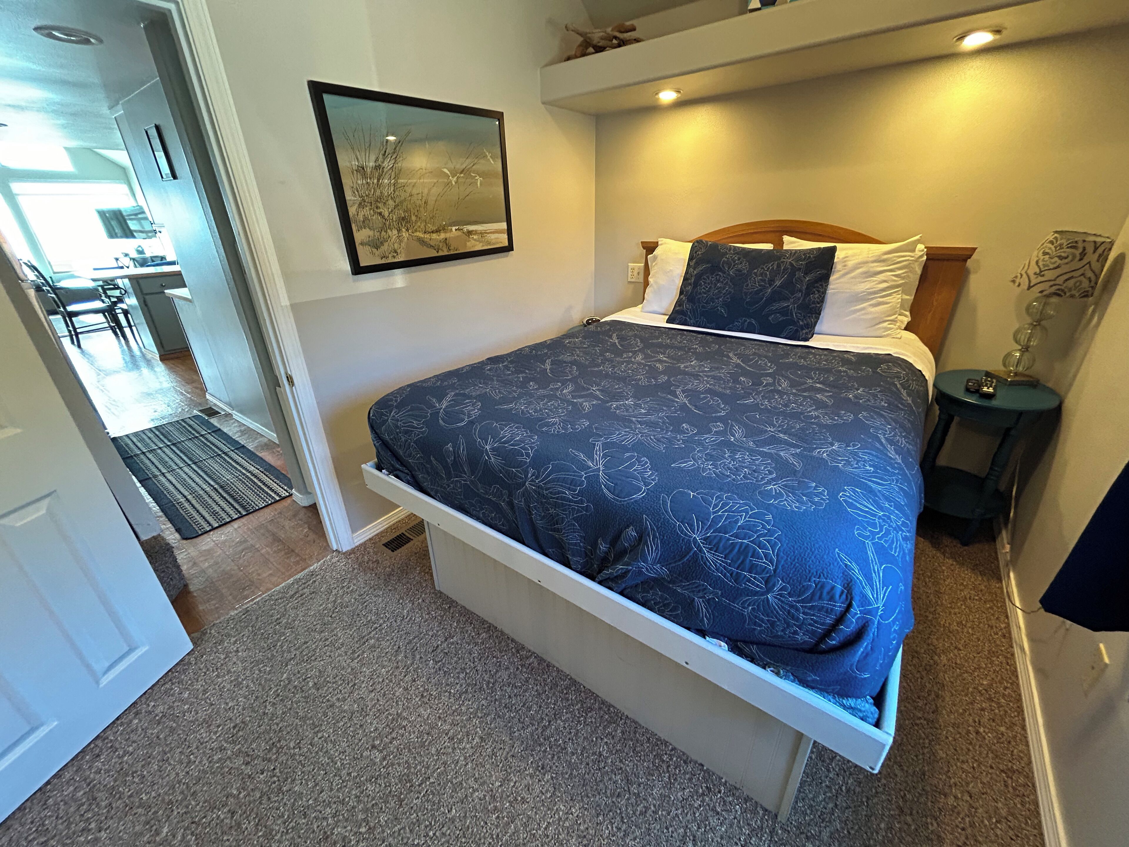 A bedroom with a bed, a nightstand, a lamp, a picture on the wall, and a door leading to a bathroom.