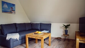 A living room with a dark blue sofa, a wooden coffee table with fruit, a small potted plant, and a television on a wooden stand. - Apartments Möwe - Apartment upper floor with lake view (Lembruch)