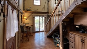 A rustic cabin interior with a wooden floor, a dining area with a table and chairs, a kitchen with cabinets and a stove, and a staircase leading to an upper level. - Amish cabin at Wolf Creek Guest Ranch (Fredericksburg)