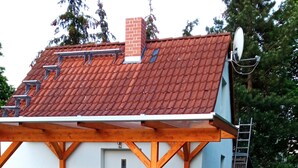 Een klein gebouw met een rode tegeltjesdak, een witte deur en een overdekt terras met metalen stoelen en een tafel.