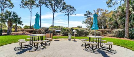 Un espace extérieur aménagé en terrasse, avec des tables de pique-nique et des parasols, entouré d’arbres et de verdure.