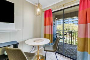 A modern room with a round table, two chairs, a flat-screen TV mounted on the wall, and a balcony with a view of buildings and greenery.