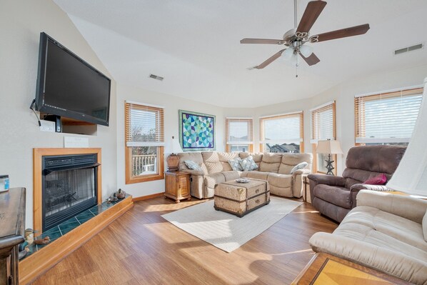 A spacious living room with a fireplace, comfortable seating, large windows, and a ceiling fan.