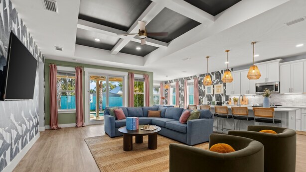 A modern living room with a sectional sofa, a coffee table, and ottomans. There is a kitchen area with white cabinets and a microwave. - Resort Pool Oasis • Game Room • Walk to Beach • Sleeps 15 (Panama City Beach)