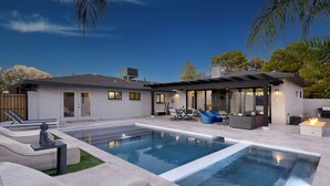 A modern house with a large rectangular pool, outdoor seating area, and a patio with a fire feature.