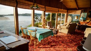 A cozy room with a large window overlooking a body of water, a dining table with chairs, and a sofa. - Panoramic Promontory: Bay View Beach House (Waldport)