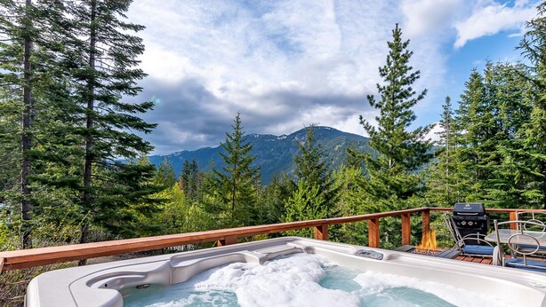 Un bain à remous avec une terrasse en bois, entouré d’arbres et avec des montagnes en arrière-plan.
