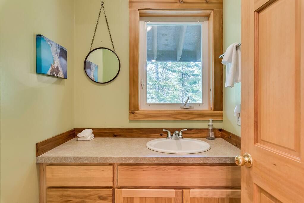 Un baño con un tocador de madera, un espejo redondo, una ventana con vistas a árboles y un perchero.
